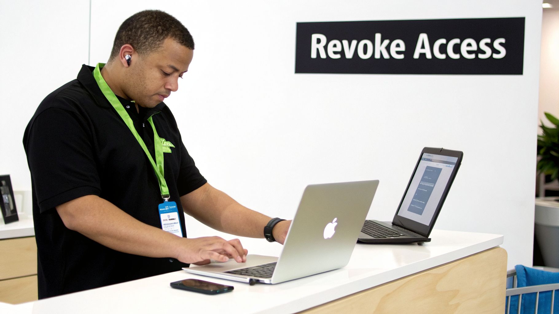 An IT professional in a black shirt works on a laptop at a counter with a 'Revoke Access' sign.