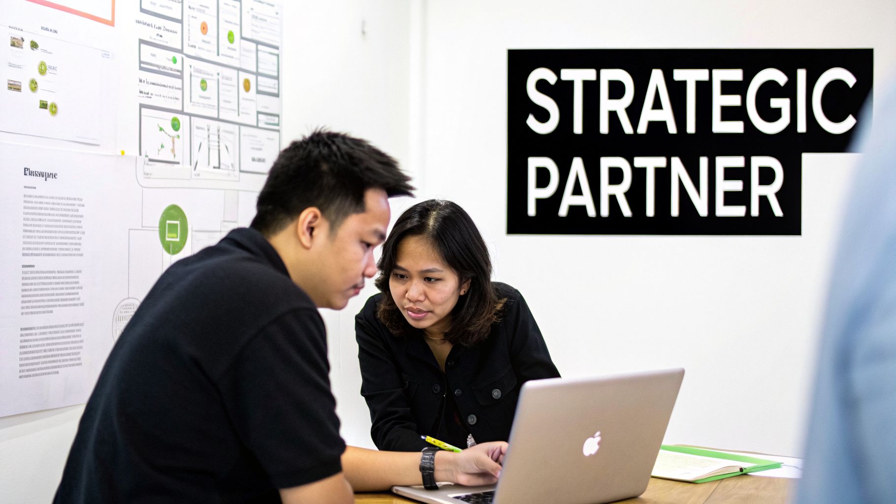 Two Asian professionals collaborate, looking at a laptop in an office with a 'STRATEGIC PARTNER' sign.